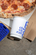 Paper coffee cup mockup tilted on a concrete dining table beside pizza.