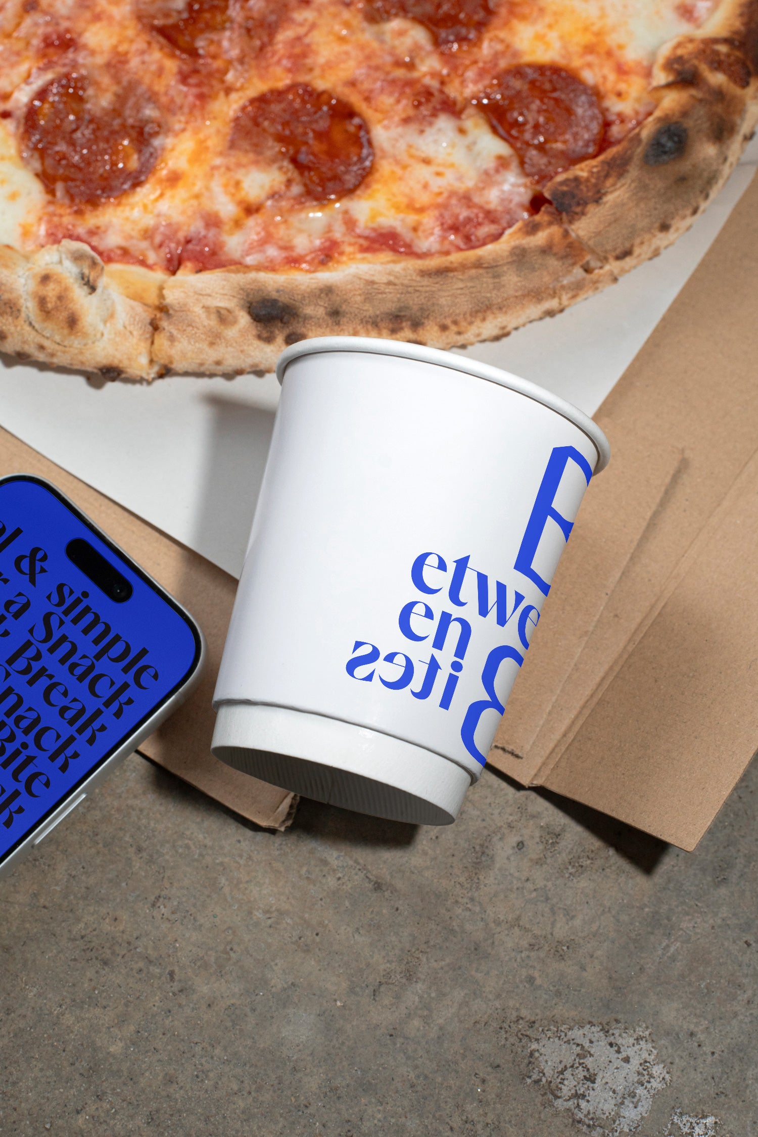Paper coffee cup mockup tilted on a concrete dining table beside pizza.