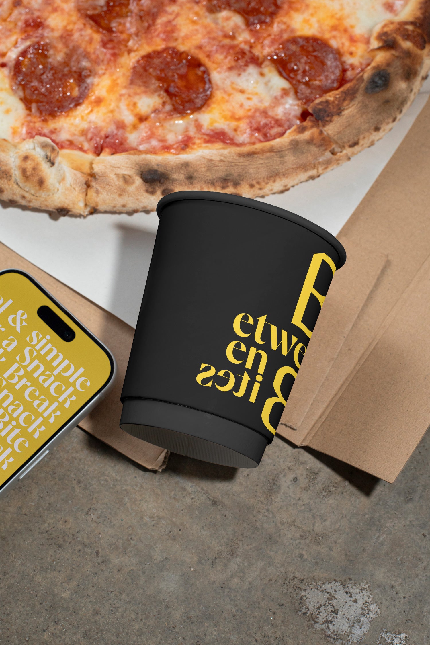 Paper coffee cup mockup tilted on a concrete dining table beside pizza.