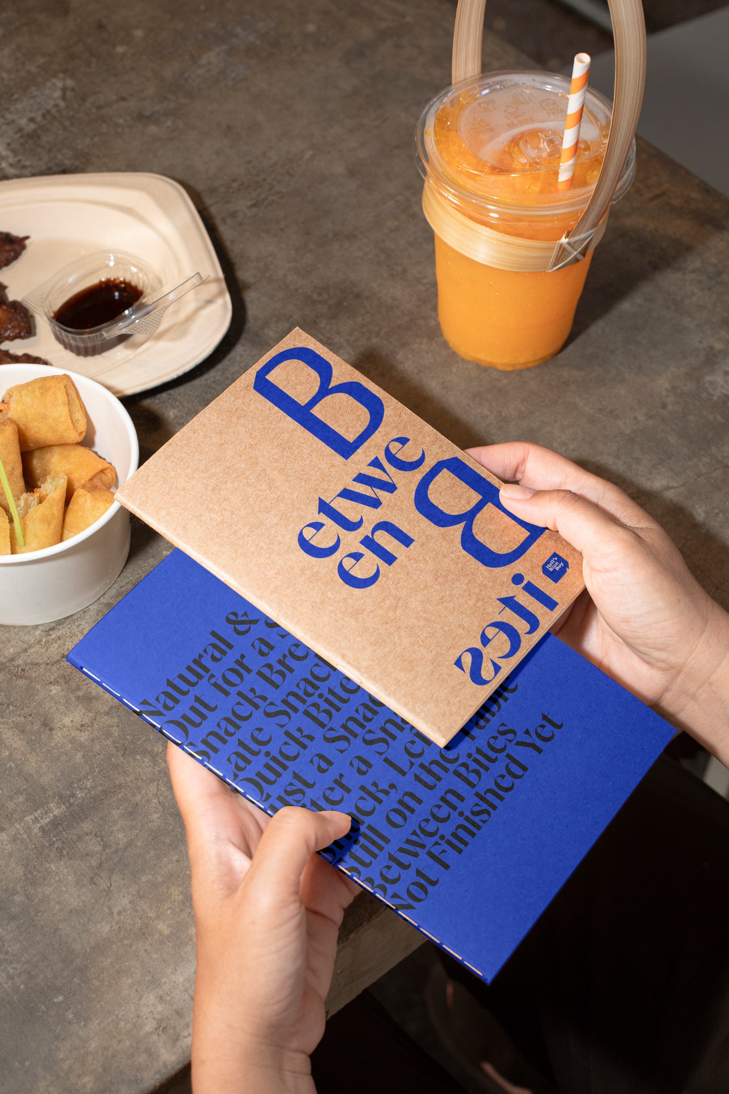 Two sketchbooks held in hand above a concrete dining table with spring rolls, grilled meat, and orange juice in the background.