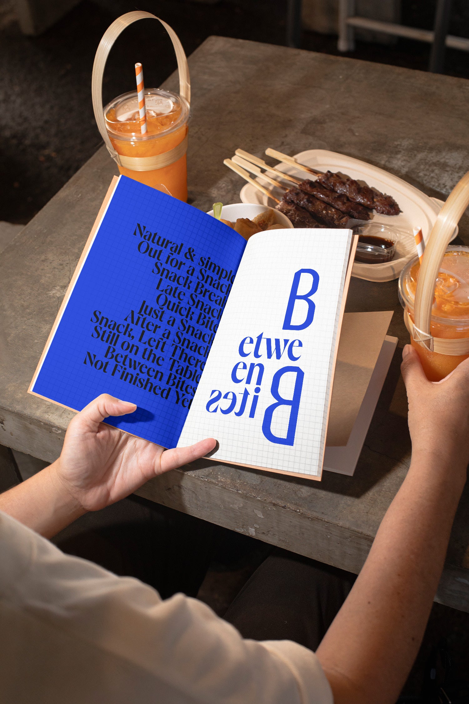 Sketchbook mockup held in hand above a concrete table with spring rolls, grilled meat, and orange juice.