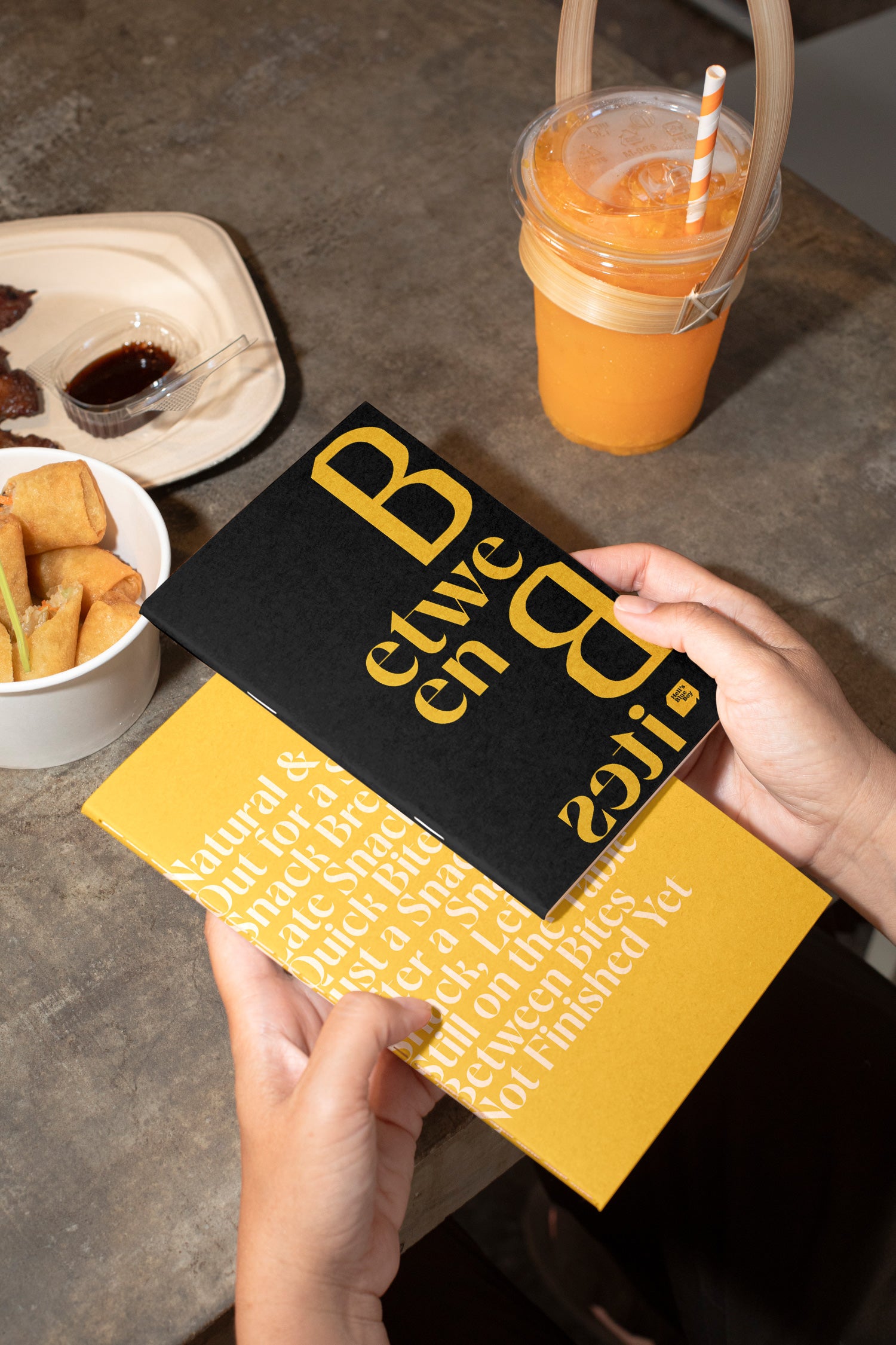 Two sketchbooks held in hand above a concrete dining table with spring rolls, grilled meat, and orange juice in the background.