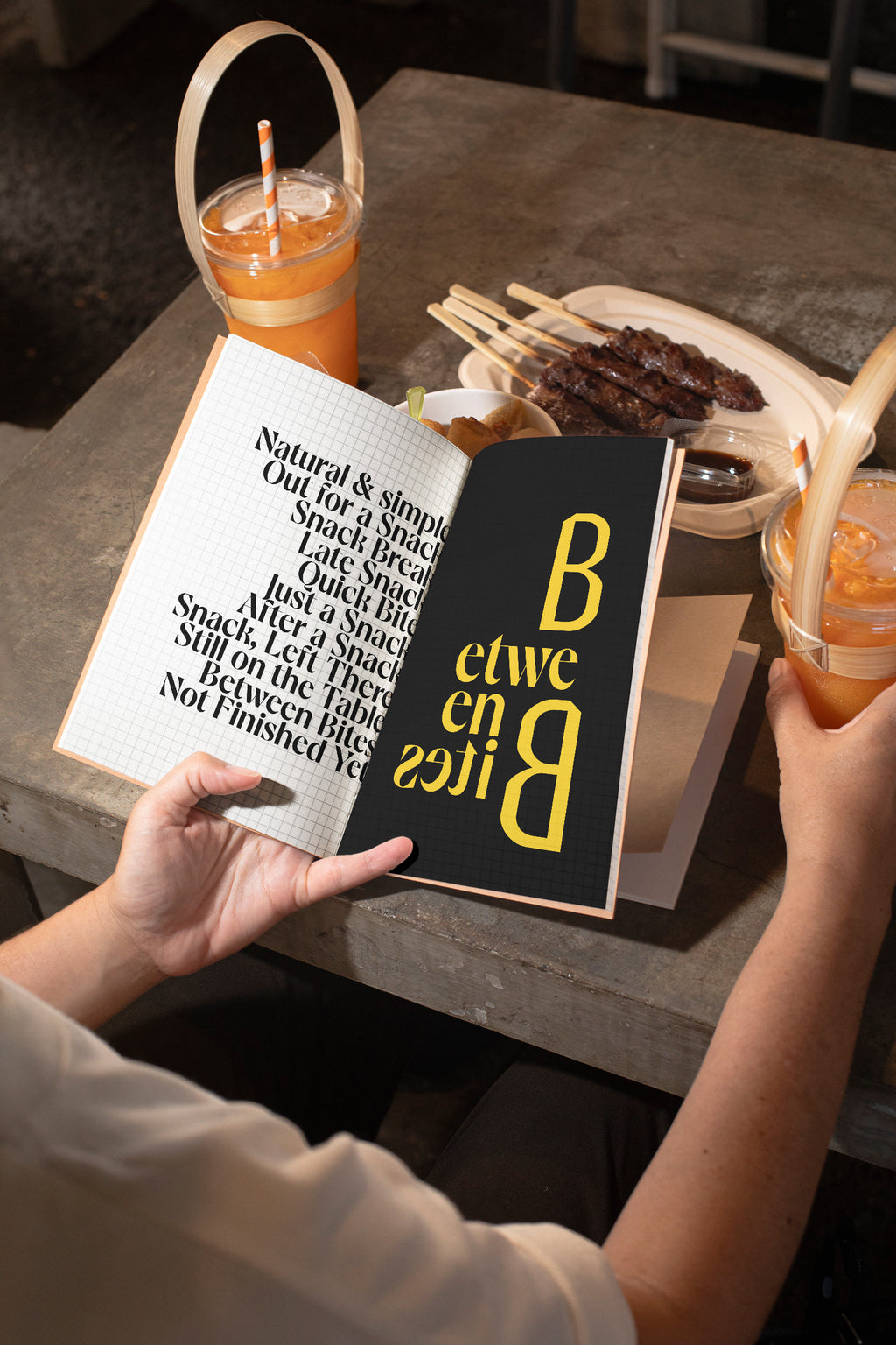 Sketchbook mockup held in hand above a concrete table with spring rolls, grilled meat, and orange juice.