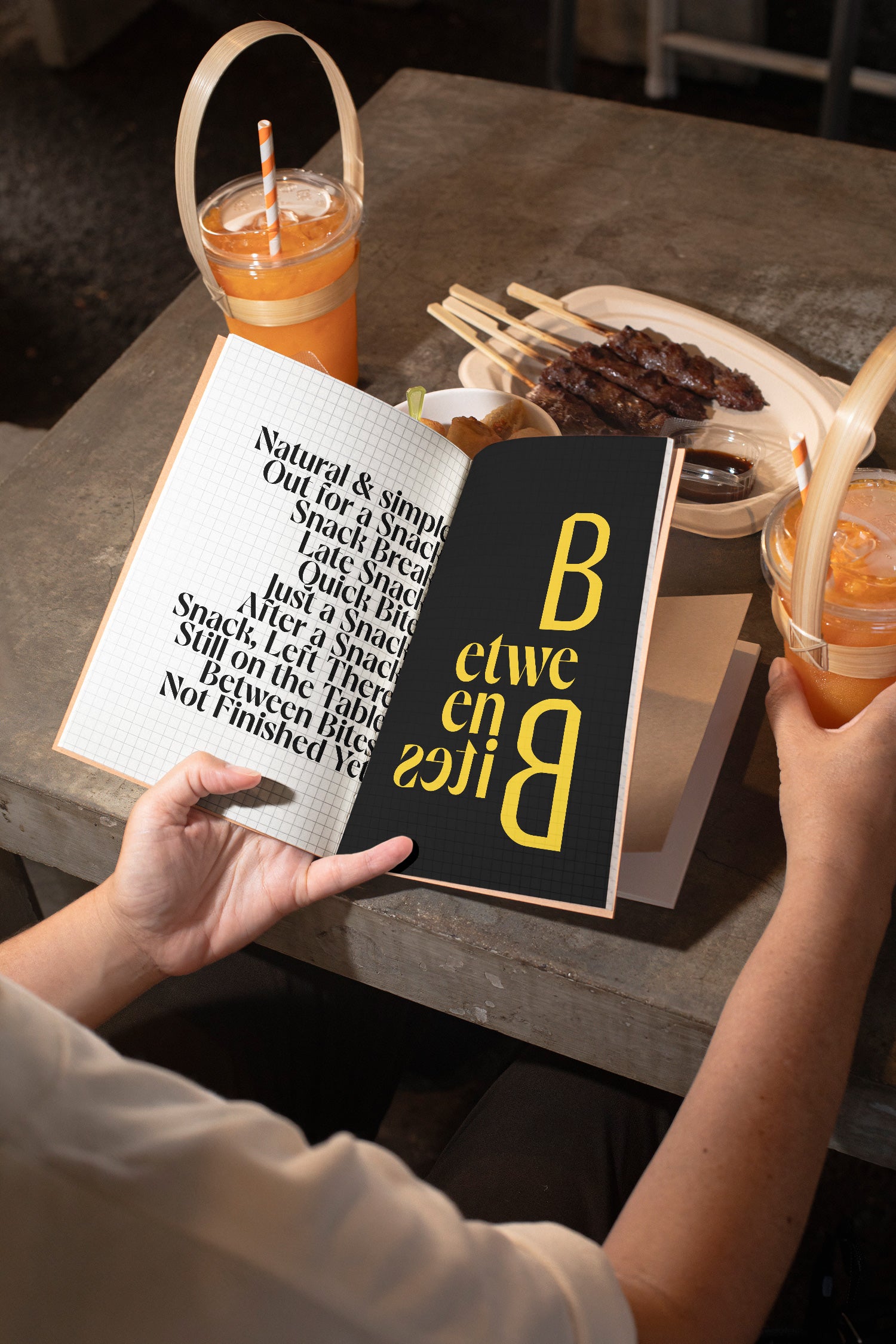 Sketchbook mockup held in hand above a concrete table with spring rolls, grilled meat, and orange juice.