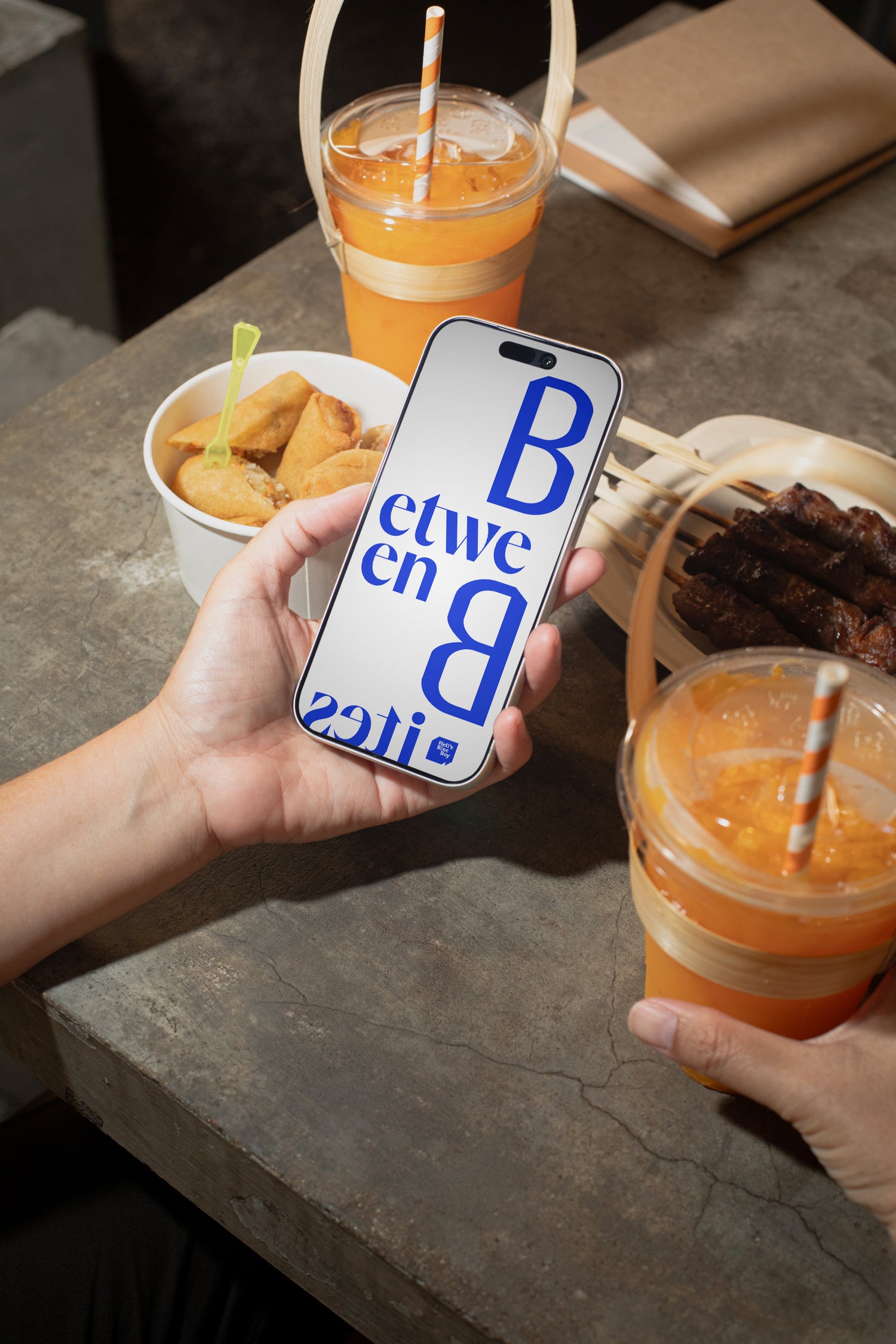 iPhone mockup held in hand above a raw concrete dining table with grilled meat, spring rolls, and an iced orange soft drink.