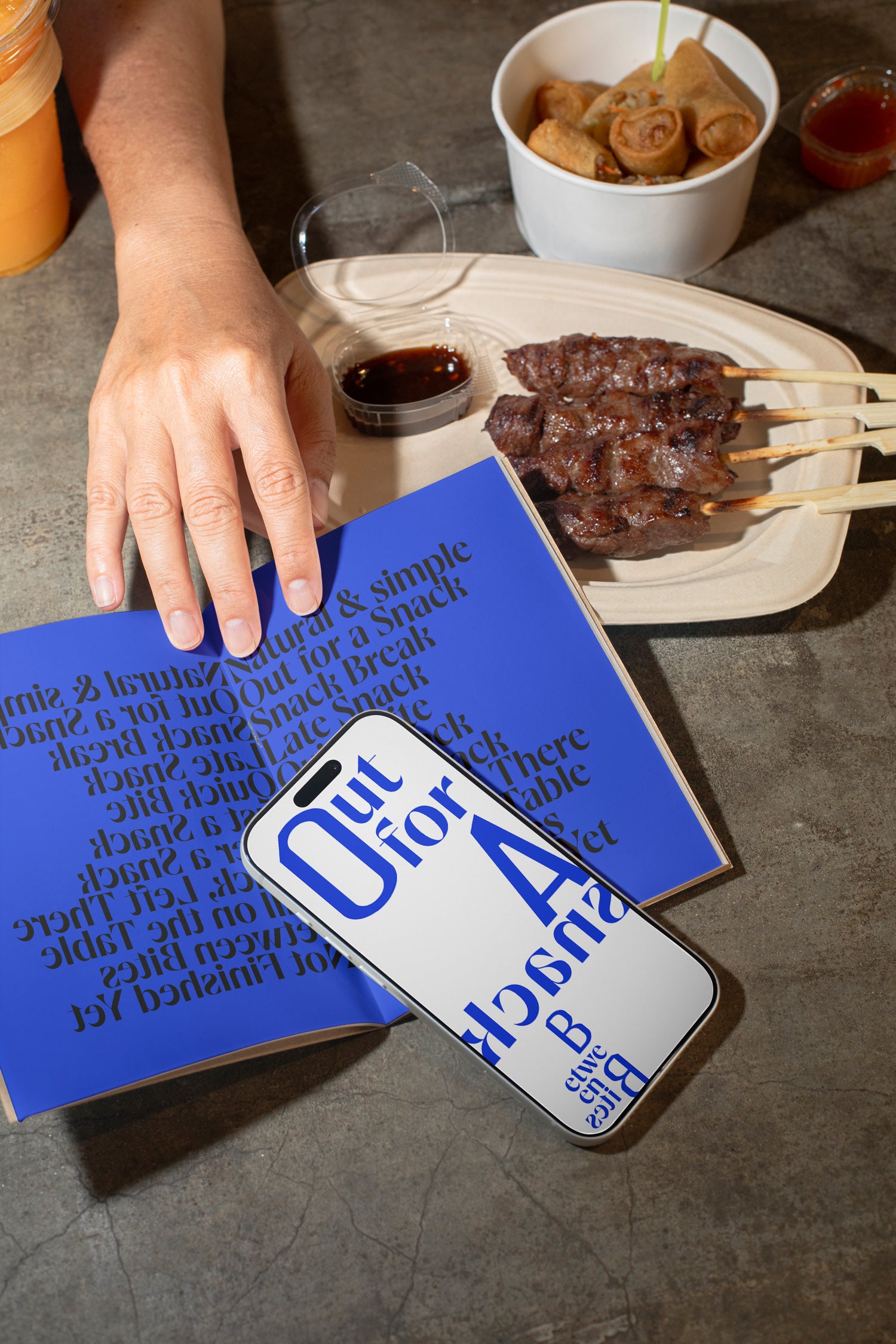iPhone mockup resting on a sketchbook with a backdrop of grilled meat, spring rolls, and a raw concrete dining table.