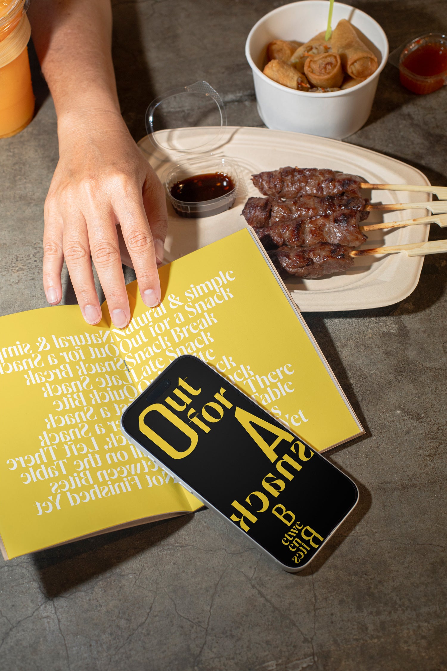 iPhone mockup resting on a sketchbook with a backdrop of grilled meat, spring rolls, and a raw concrete dining table.