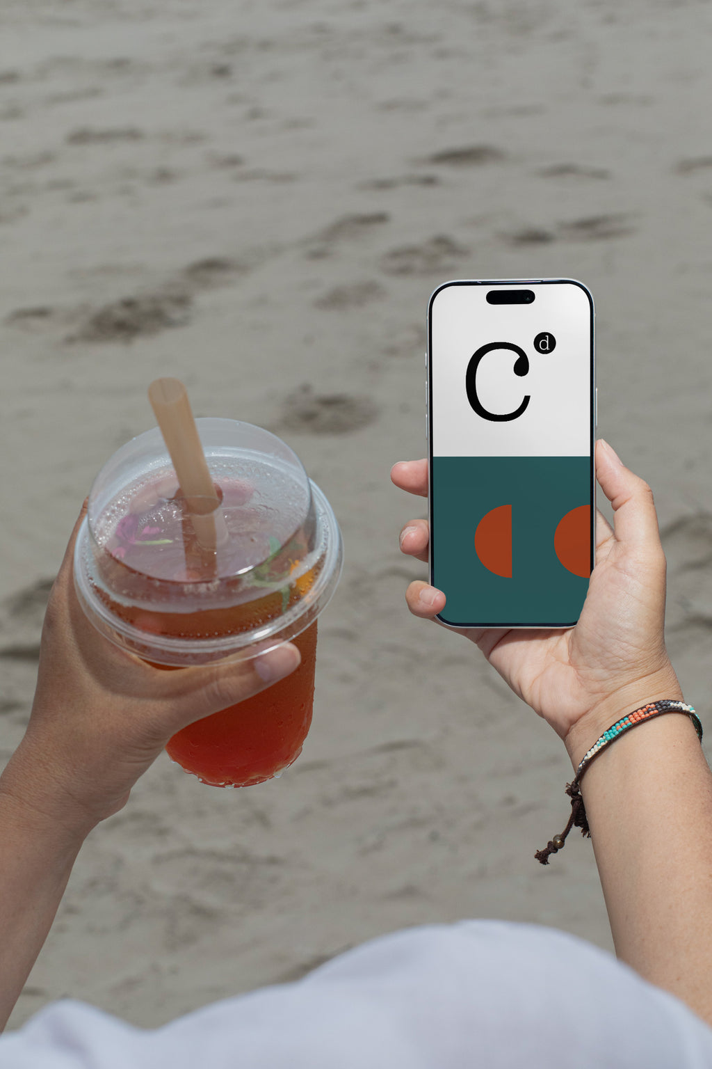 iPhone mockup held by hand with soda cup against a sandy beach backdrop. Built for showcasing app interfaces, branding, editorial layouts, illustrations, and creative presentations.