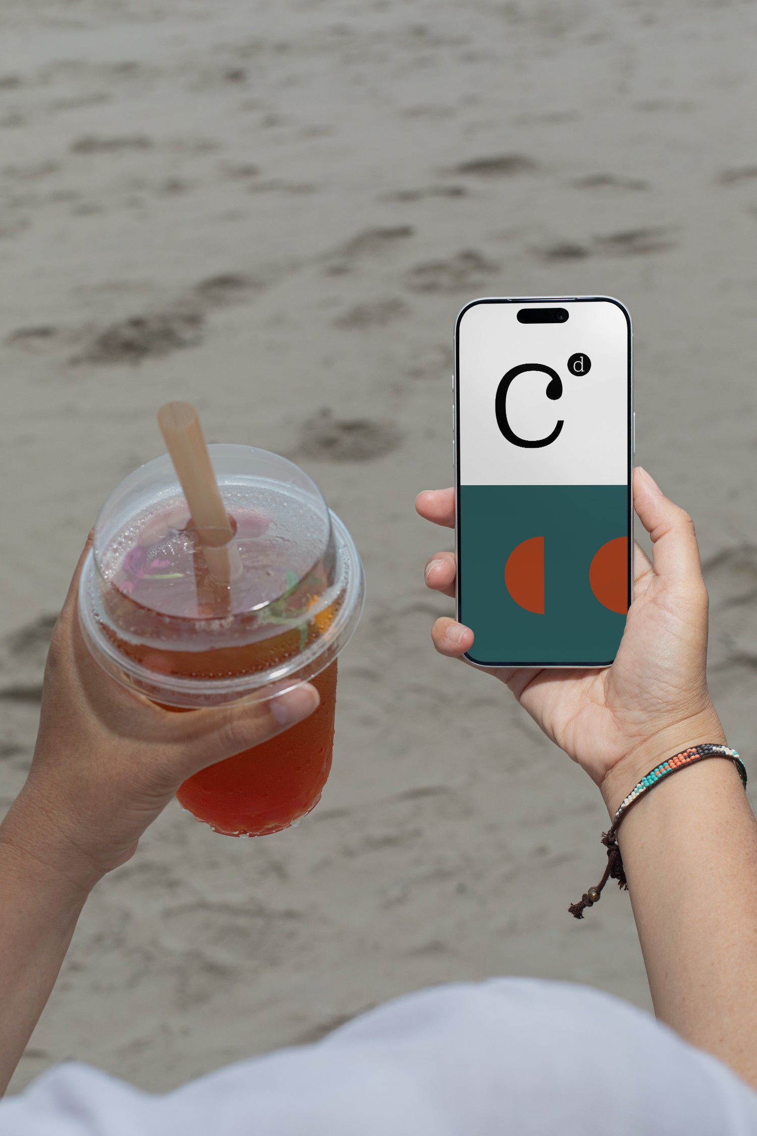 iPhone mockup held by hand with soda cup against a sandy beach backdrop. Built for showcasing app interfaces, branding, editorial layouts, illustrations, and creative presentations.