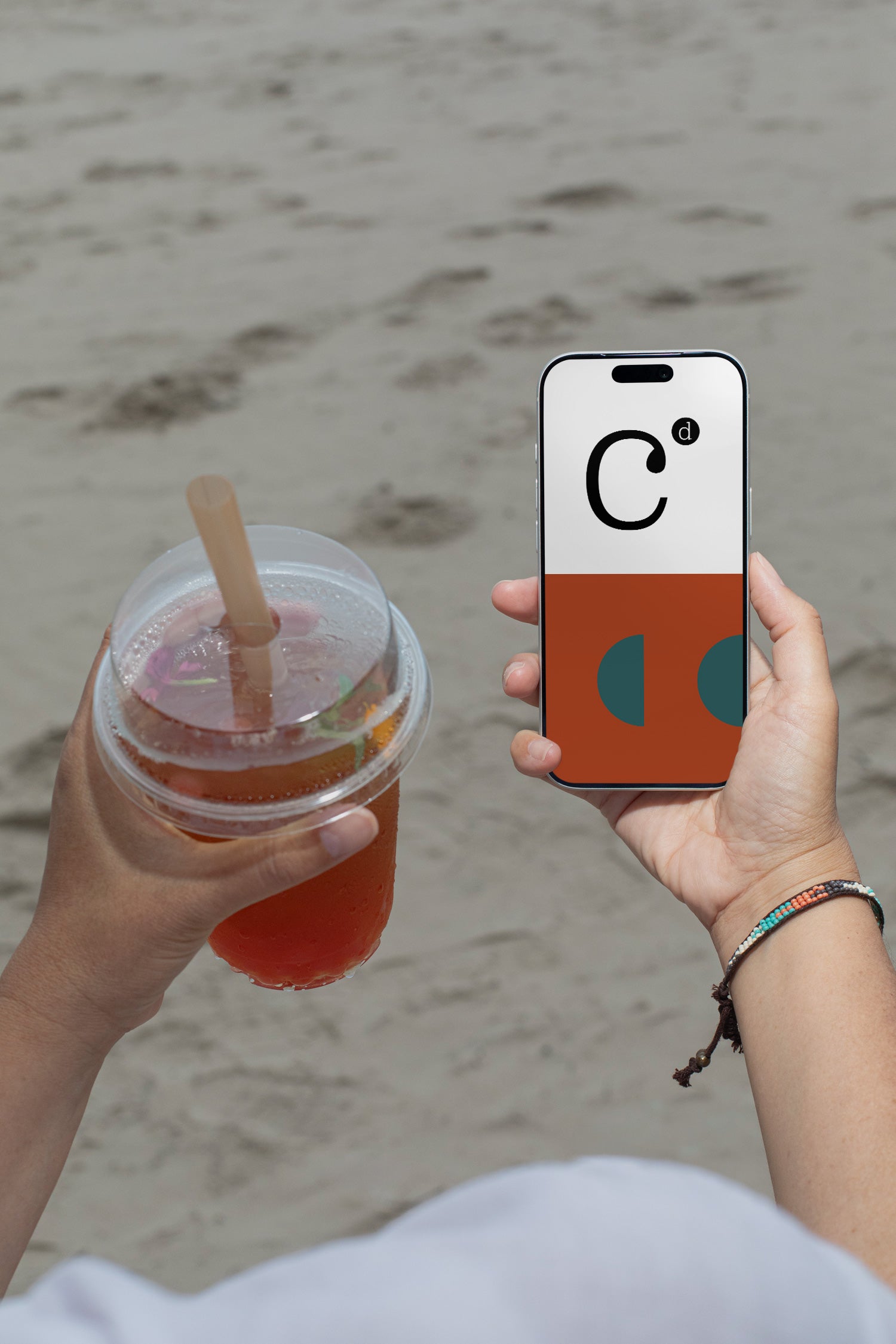 iPhone mockup held by hand with soda cup against a sandy beach backdrop. Built for showcasing app interfaces, branding, editorial layouts, illustrations, and creative presentations.