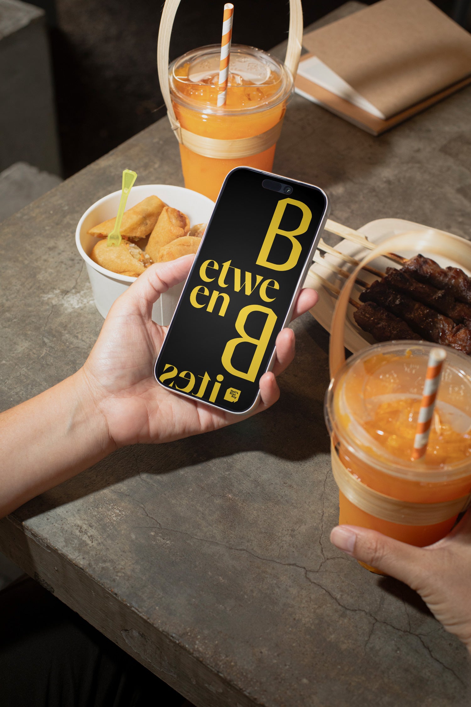 iPhone mockup held in hand above a raw concrete dining table with grilled meat, spring rolls, and an iced orange soft drink.