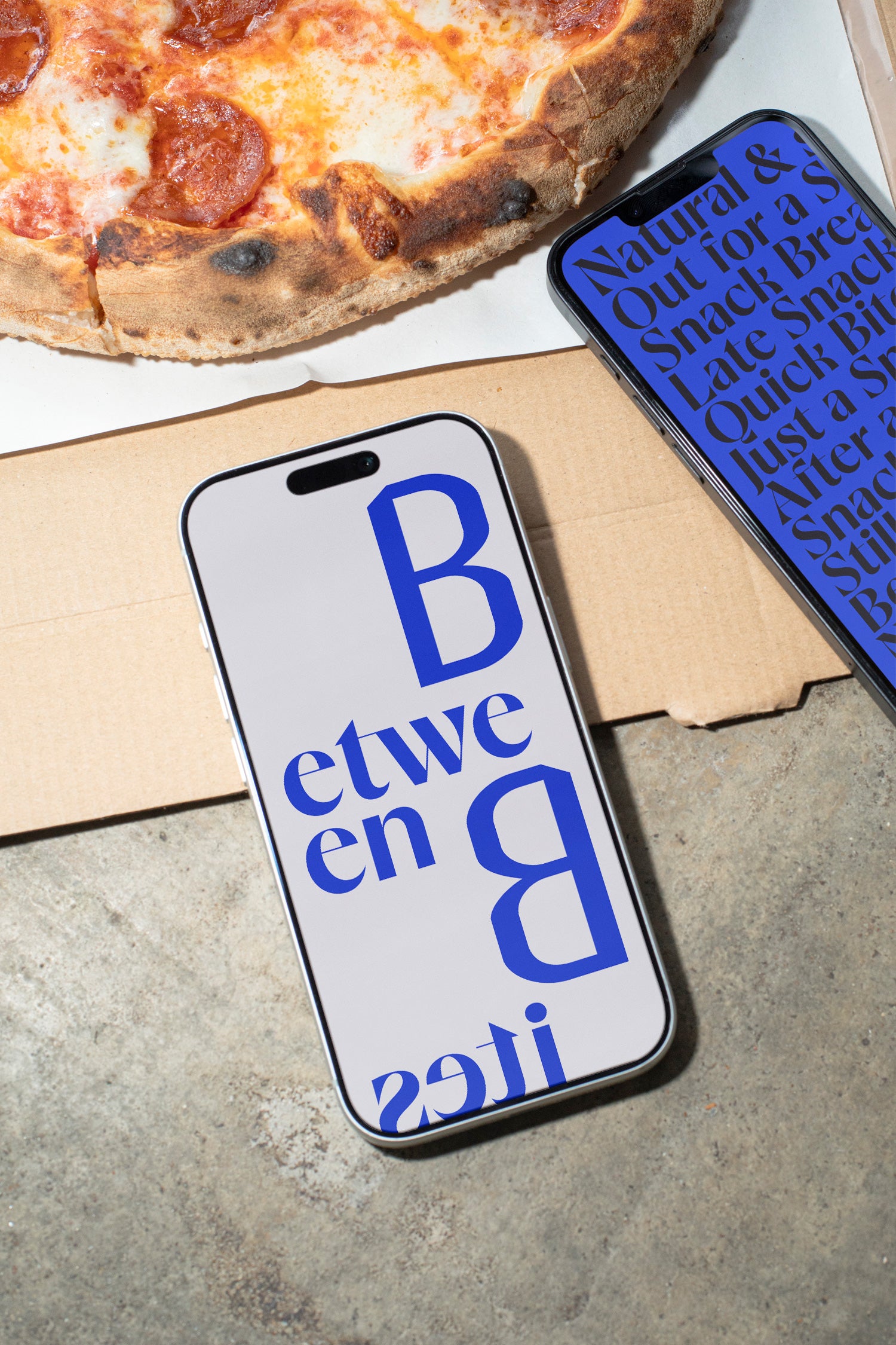 Two iPhones lying on a concrete table beside pizza and a takeaway box.