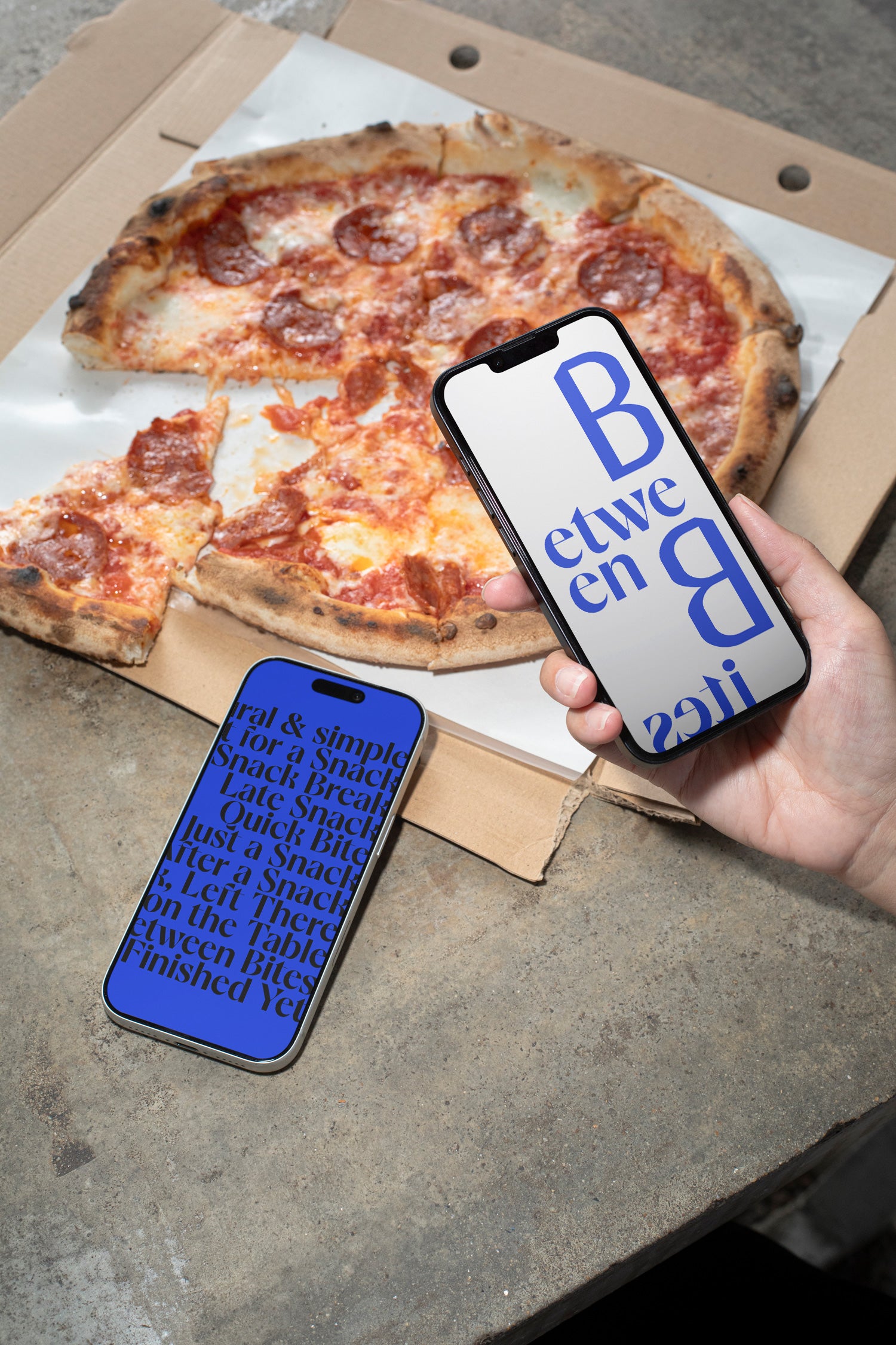 Two iPhones lying on a concrete table beside pizza and a takeaway box.
