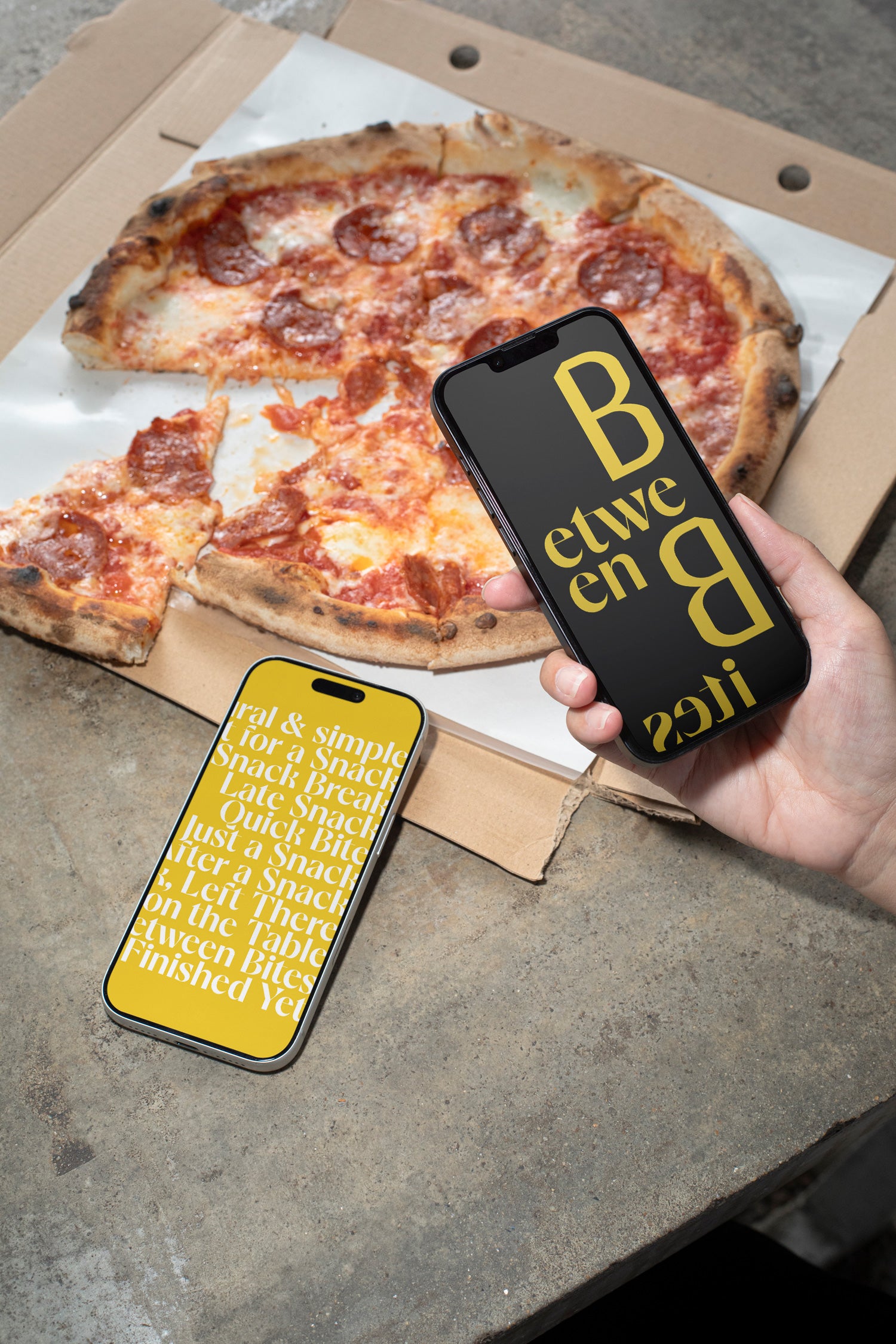 Two iPhones lying on a concrete table beside pizza and a takeaway box.